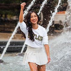 a young woman is standing in front of a water fountain and raising her hand up