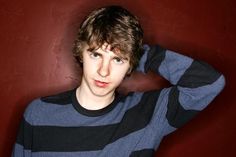 a young man poses in front of a red wall with his hand on his head