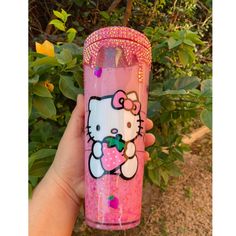 a hand holding a hello kitty tumbler in front of some bushes and flowers,