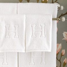 two white towels with the letter h on them sitting next to each other in front of a floral wallpaper