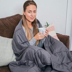 a woman sitting on a couch holding a coffee cup and blanket over her head, while wrapped in a blanket