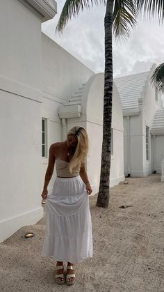 a woman in a white dress standing next to a palm tree on the sand near a building