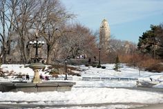 a fountain in the middle of a snowy park with people walking around it and trees