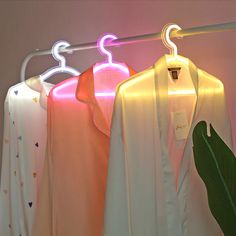 three shirts hanging on a clothes rack with neon lights in the corner and a green plant next to them