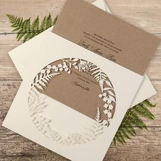 two wedding cards on top of each other with leaves and ferns in the middle, sitting on a wooden table