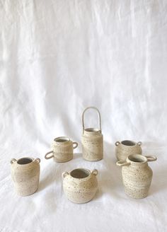 four white ceramic pots with handles on a white cloth covered tablecloth, set against a plain backdrop