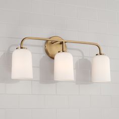 three light bathroom fixture with white glass shades on the sides and gold finish, against a white brick wall