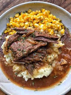 a white plate topped with meat and mashed potatoes covered in gravy next to corn