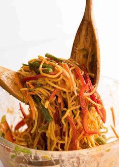 a wooden spoon filled with noodles and veggies in a glass bowl on top of a table