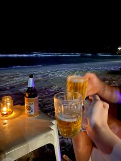 two people toasting with beer on the beach in front of an ocean at night