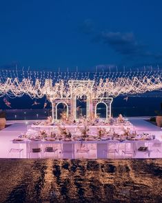 an outdoor dinner table set up with white linens and lights on the ceiling above it