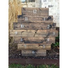 a clock made out of wooden pallets sitting on top of some grass next to a brick wall