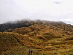 two people walking up the side of a mountain on a cloudy day with mountains in the background