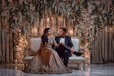 a man and woman sitting on top of a couch in front of a floral wall