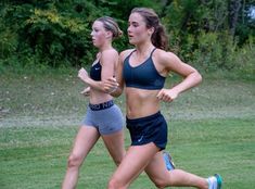 two women are running on the grass in their sports bras and shorts, with trees in the background