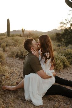 a man and woman sitting on the ground kissing in front of a cactus field at sunset