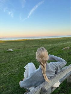 a woman sitting on top of a wooden bench next to the ocean with her hair in a braid