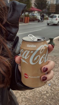 a woman holding up a can of coca - cola in front of a street with parked cars