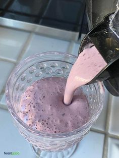 a person pours pink liquid into a glass cup on a tile counter top with a blender in the background