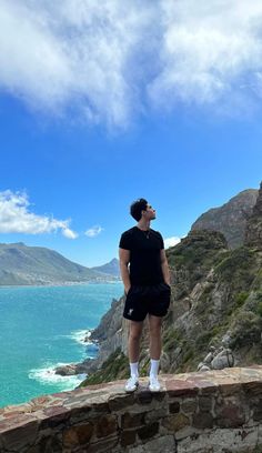 a man standing on top of a stone wall next to the ocean with mountains in the background