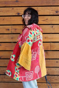 a woman standing in front of a wooden wall wearing a red and yellow jacket with geometric designs on it