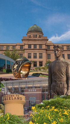 a statue of a man in front of a large building with flowers and plants around it