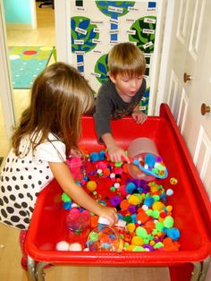 two young children playing with toys in a red plastic bin on the floor next to a white door