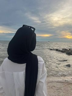 a woman wearing a white shirt and black scarf standing on the beach looking out at the ocean