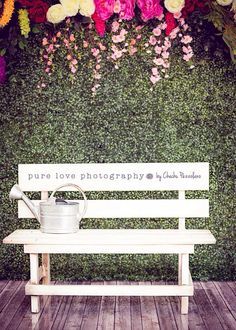 a wooden bench sitting in front of a green wall with flowers and a watering can