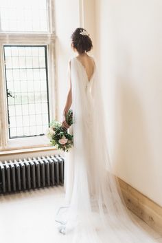 a woman in a wedding dress standing by a window with her back to the camera