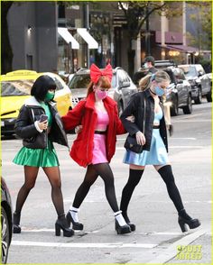 three girls in costumes crossing the street with their hands on their hipss and wearing masks