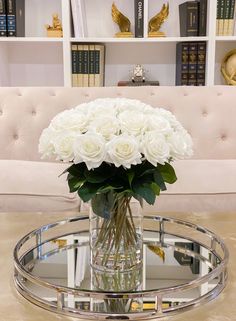 a vase with white roses on a table in front of a couch and bookshelves