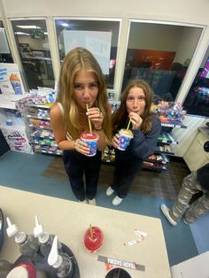 two girls are standing in front of a store counter drinking sodas and eating donuts