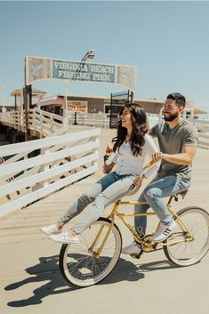 a man and woman riding a bike on the beach in front of a wooden pier