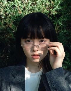 a woman wearing glasses standing in front of some bushes with her hand on her head