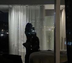 a person standing in front of a window with curtains on the windowsill and city lights behind them