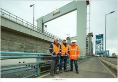 three men in orange vests standing next to each other on a bridge over water