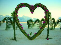 a heart made out of branches and flowers on the beach with palm trees in the background