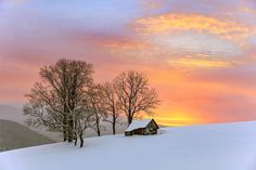 a house in the middle of a snow covered field with trees and mountains in the background