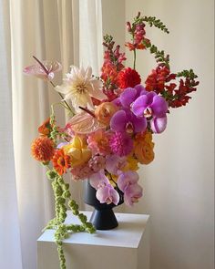 a black vase filled with lots of colorful flowers on top of a white table next to a window