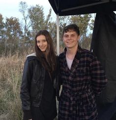 a man and woman standing next to each other in front of a black tent with trees behind them