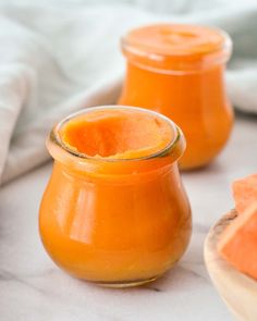 two jars filled with orange colored liquid sitting on top of a white tablecloth next to slices of watermelon