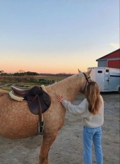 a woman standing next to a brown horse on top of a dirt field with a red barn in the background