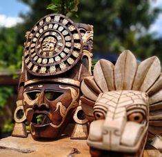 two wooden masks sitting next to each other on top of a piece of wood with trees in the background