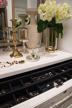 a white dresser topped with lots of jewelry next to a vase filled with flowers and candles