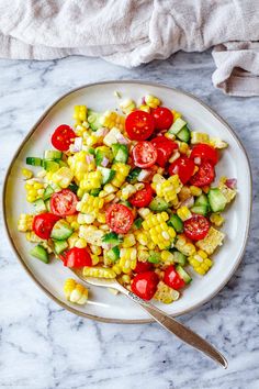 a white plate topped with corn, tomatoes and cucumbers next to a fork