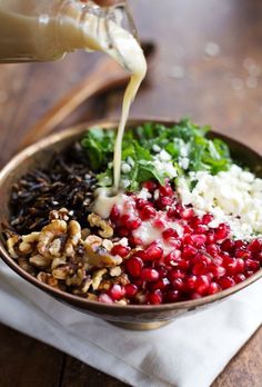 someone is pouring dressing into a bowl filled with vegetables and grains, including pomegranates