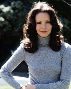 a woman with long hair wearing a gray turtle neck sweater posing for a photo in front of some trees