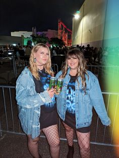 two women standing next to each other in front of a fence at night, one holding a drink and the other wearing fish net stockings