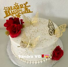 a white cake with gold butterflies and red roses on top is sitting on a table